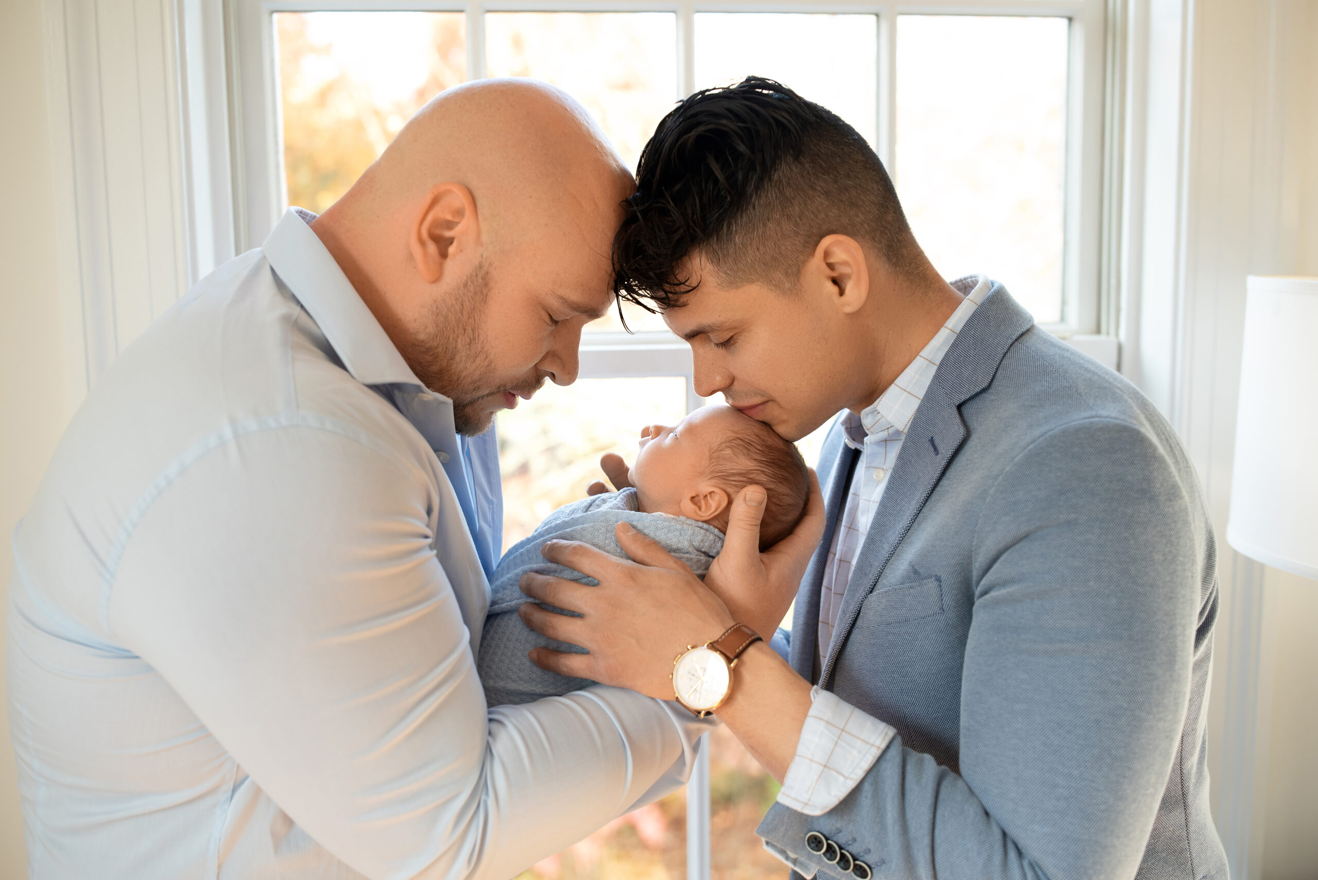 two dads holding newborn baby closely with eyes closed in front of window