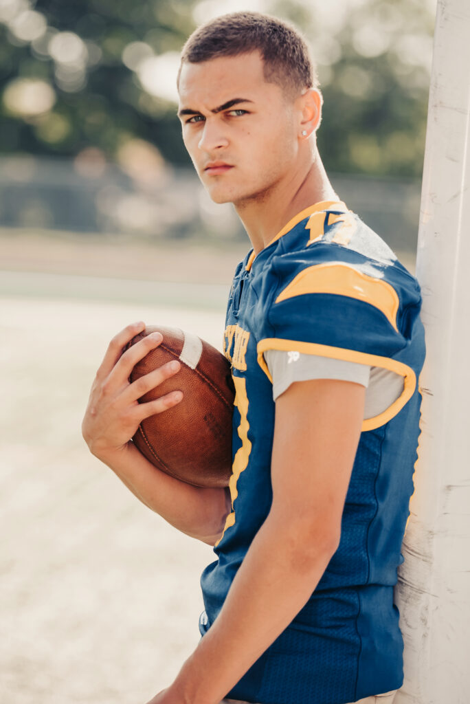 Senior guy wearing a football jersey standing against a wall and holding a football in Westport Connecticut