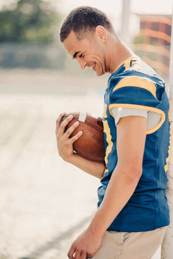 Senior guy wearing a football jersey standing against a wall and holding a football in Westport Connecticut